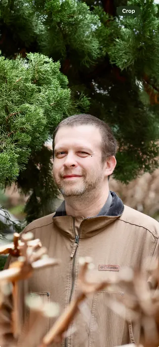 A man in a tan jacket smiling beneath evergreen branches