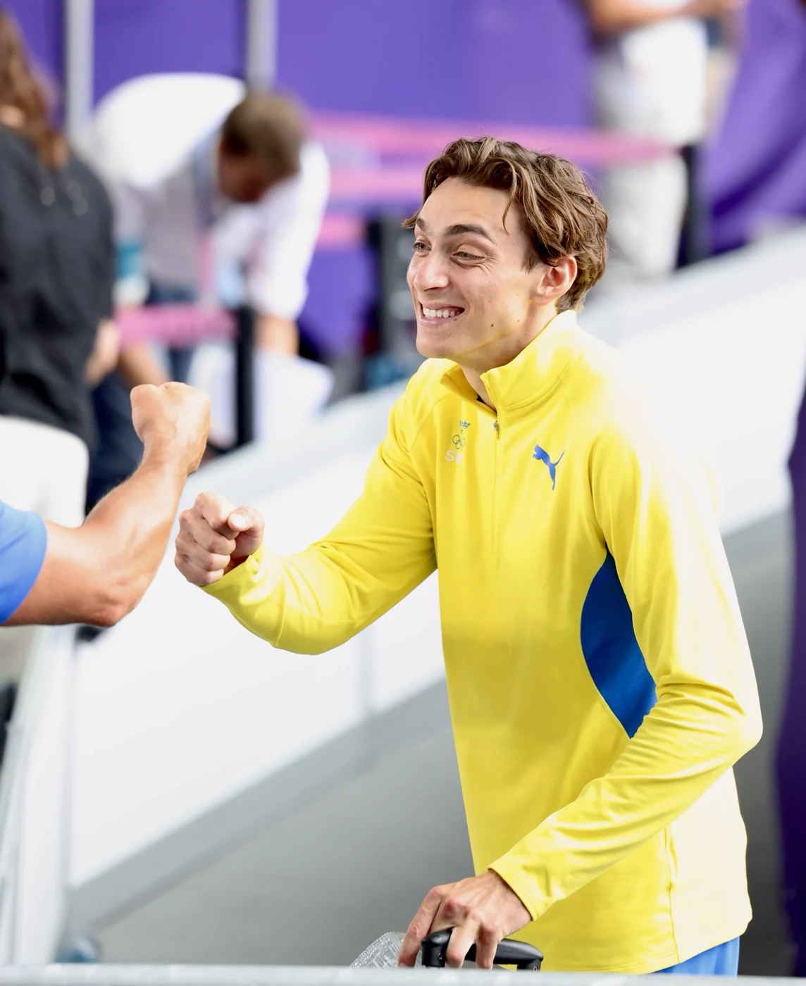 An Olympic athlete in a yellow warm-up top smiling and bumping fists at the Games